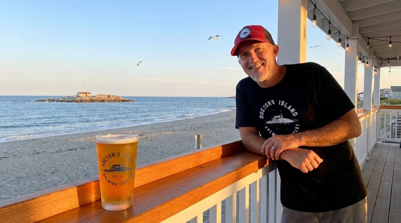 Doctor's Island Brewing owner Greg Hoffmeister on the outdoor patio in Hull, Massachusetts with a pint of amber beer and the actual Doctor's Island visible offshore